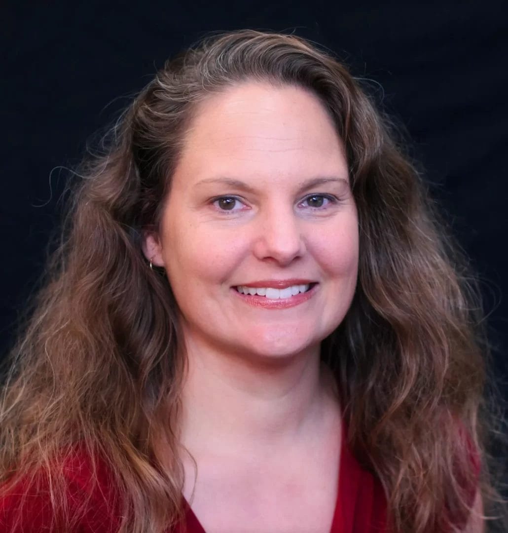 A woman with long wavy hair, wearing a red shirt, smiling at the camera against a black background.